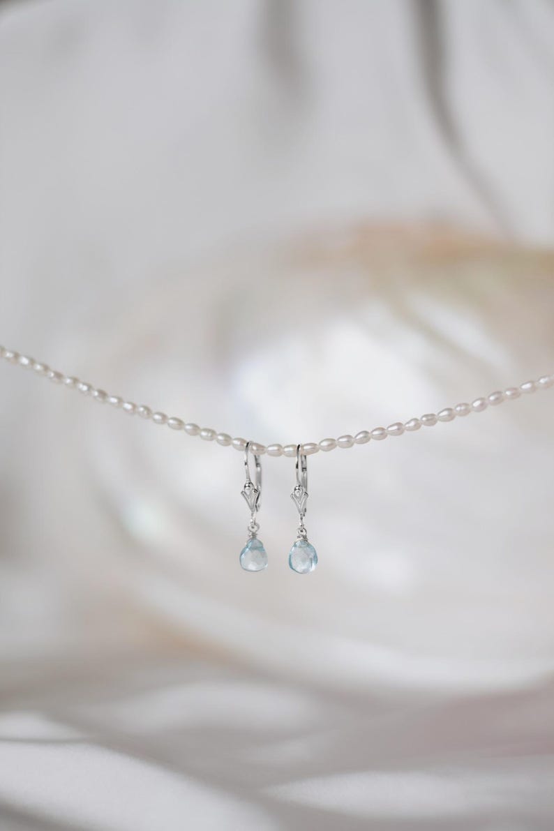 Op de afbeelding: Een paar zilveren oorhangers met lichtblauwe, druppelvormige edelstenen. De oorbellen hangen aan een delicate parelketting tegen een zachte, witte achtergrond. De oorbellen hebben een decoratief ontwerp boven de edelsteen.