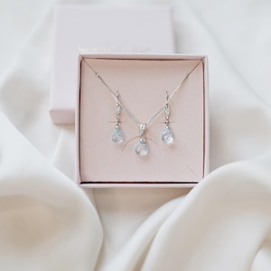 Op de afbeelding: Een zilveren ketting- en oorbellenset met druppelvormige, lichtblauwe edelstenen, gepresenteerd in een roze sieradendoosje. De set bevat een hangerketting en bijpassende oorhangers. Het doosje is open en toont de sieraden tegen een zachte, witte stoffen achtergrond.