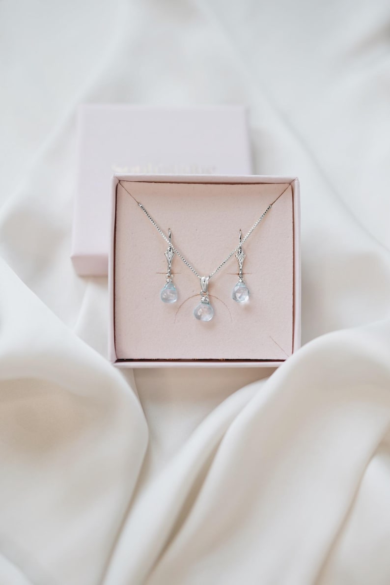 Op de afbeelding: Een zilveren ketting- en oorbellenset met druppelvormige, lichtblauwe edelstenen, gepresenteerd in een roze sieradendoosje. De set bevat een hangerketting en bijpassende oorhangers. Het doosje is open en toont de sieraden tegen een zachte, witte stoffen achtergrond.