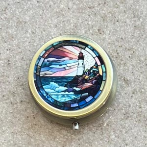 May include: Round, brass-colored pill box with a stained-glass design of a lighthouse on a rocky coast. The artwork features a colorful sunset, blue waves, and a white lighthouse. The box has a hinged lid and a small clasp.