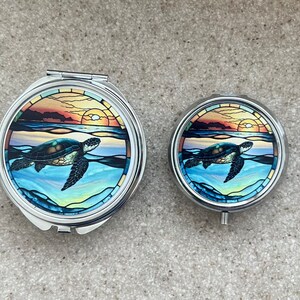 May include: Two silver compact mirrors with a stained glass design of a sea turtle swimming in the ocean at sunset. The larger mirror is slightly larger than the smaller mirror.