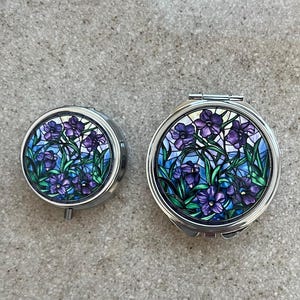 May include: Two round silver compact mirrors with a floral design. The larger mirror has a stained glass look with purple flowers and green leaves. The smaller mirror has a similar design but is more stylized.