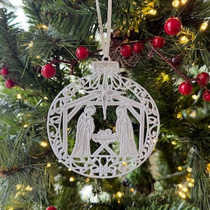 May include: A white, intricately designed Christmas ornament depicting the Nativity scene. The ornament is round with a detailed lace-like pattern, hanging from a white ribbon. It is set against a backdrop of a Christmas tree with red berries and lights.