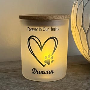 May include: A frosted glass candle jar with a bamboo lid. The jar has a black paw print inside a heart with the text "Forever In Our Hearts" and "Duncan" below the heart.