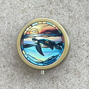 May include: A round metal pill box with a stained glass design of a sea turtle swimming in the ocean at sunset. The turtle is blue and green, and the ocean is blue and green with a yellow sunset in the background.