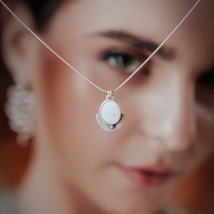 Puede incluir: Un collar de plata con un colgante ovalado blanco y un diseño de media luna. El colgante cuelga frente al rostro de una mujer, que está desenfocado.