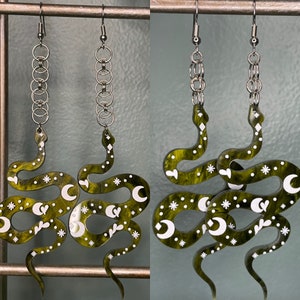 Op de afbeelding: Een paar groene slangenoorbellen met witte halve manen en sterren. De oorbellen zijn gemaakt van hars en hebben een zilveren ketting.