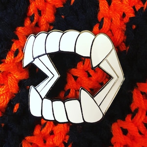 May include: White enamel pin shaped like a set of fangs. The pin is on a black and orange crocheted background.