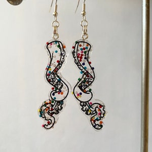 Könnte beinhalten: Klare Acryl-Ohrringe in Form von stilisierten Tentakeln, schwarz umrandet, mit bunten, konfettiartigen Akzenten. Die Ohrringe hängen an silberfarbenen Fischhaken-Ohrdrähten. Das Design ist skurril und verspielt.