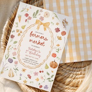 May include: A rectangular invitation card with a white background featuring a watercolour floral and pumpkin border, a gold bow, and the text farmers market to celebrate Katelyn's 1st Birthday, paired with a matching yellow and white gingham envelope.