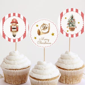 Op de afbeelding: Drie cupcakes met witte glazuur, elk bekroond met een decoratie met kerstthema. De toppers tonen een notenkraker, een Franse hoorn en een kerstboom, alles tegen een rood-witte gestreepte achtergrond. De middelste topper zegt "Merry Christmas".