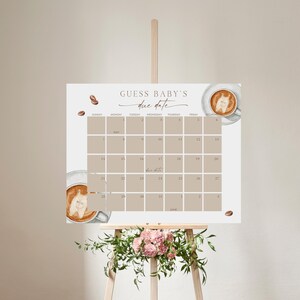 May include: A baby shower calendar with the text "Guess Baby's Due Date" in elegant script. The calendar features a neutral color palette with coffee cup illustrations and coffee beans. The calendar is displayed on a wooden easel.
