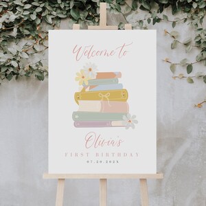 May include: Welcome to Olivia's First Birthday sign with a stack of colorful books and flowers. The date 07.20.202X is printed below the text.