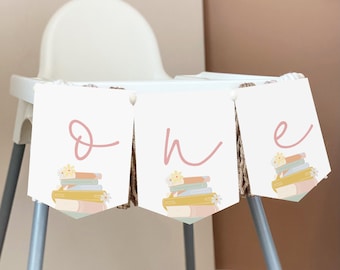 May include: A white banner with pink lettering that reads "one" with a stack of books and flowers on each letter. The banner is strung on a high chair.