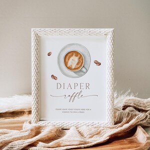 May include: A framed sign with a watercolor illustration of a coffee cup and beans. The sign reads "DIAPER raffle" with additional text below. The frame is white with a textured design. The sign is on a wooden tray with fabric.
