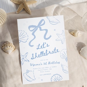 May include: A white invitation card with blue seashell illustrations and the text "Let's Shellebrate" for a first birthday. The card is surrounded by seashells and starfish on a beige fabric.