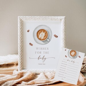 May include: A framed print with a coffee cup illustration and the text "Wishes for the baby". A matching card with a coffee cup illustration and fill-in-the-blank prompts is also present. Coffee beans are scattered around the frame.