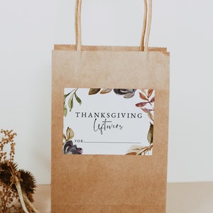 Op de afbeelding: Een bruine papieren tas met een wit label waarop "Thanksgiving Leftovers for..." staat met een aquarelbloemendesign. De tas heeft twee bruine papieren handvatten.