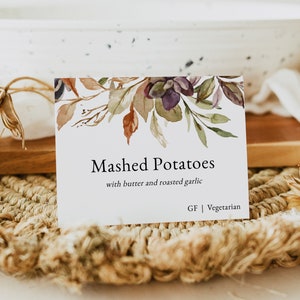 May include: A white card with a watercolor floral design in shades of purple, brown, and green. The card reads "Mashed Potatoes with butter and roasted garlic. GF | Vegetarian."  The card is sitting on a woven placemat.