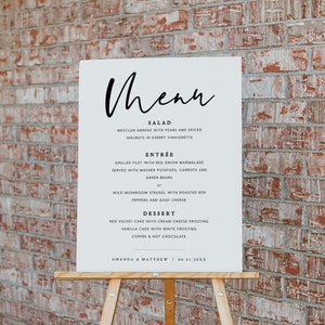 May include: A white sign with black text on an easel, listing the menu for a wedding reception. The menu includes salad, entree, dessert, and coffee and hot chocolate. The sign is titled "Menu" and includes the names Amanda & Matthew and the date 04.21.20XX.