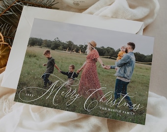 May include: A Christmas card featuring a family of four standing in a field. The card has a white background with a glittery effect and the text "Merry Christmas" in cursive. The text "The Martin Family" is printed below the greeting.