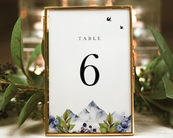 Op de afbeelding: Een gouden geframed tafelnummerkaart met een witte achtergrond. Het nummer "6" is in zwart gedrukt, met het woord "TABLE" erboven. De onderkant van de kaart heeft een aquarel-illustratie van een bergketen met blauwe bessen en bladeren.