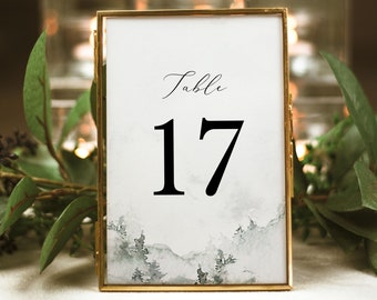 Op de afbeelding: Een tafelnummerkaart met een gouden frame en een aquarel achtergrond van een bos scène. De kaart heeft de tekst "Tafel 17" in zwarte letters.