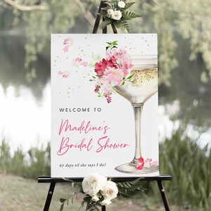 May include: A white welcome sign with pink floral accents and a champagne glass illustration. The text reads "Welcome to Madeline's Bridal Shower 67 days til she says I do!"