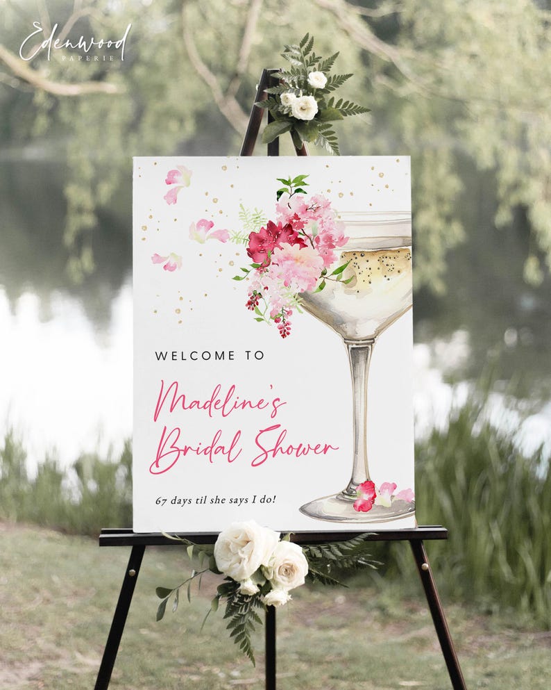 May include: A white welcome sign with pink floral accents and a champagne glass illustration. The text reads "Welcome to Madeline's Bridal Shower 67 days til she says I do!"