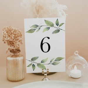 May include: White card with a green watercolor leaf design and the number 6 printed in black. The card is on a gold metal stand and is sitting on a table with a white tablecloth.
