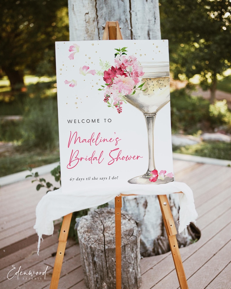 May include: A white sign with gold glitter and pink flowers announcing a bridal shower for Madeline. The sign says "Welcome to Madeline's Bridal Shower 67 days til she says I do!"