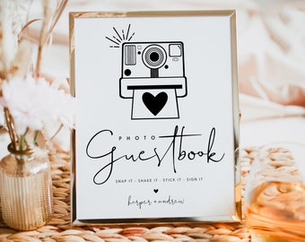 May include: A white sign with a gold frame, featuring a black and white illustration of an instant camera with a heart coming out of it. The text "PHOTO GUESTBOOK" is written in black cursive font. Below the text, it says "SNAP IT - SHAKE IT - STICK IT - SIGN IT" and "harper + andrew" in black font.
