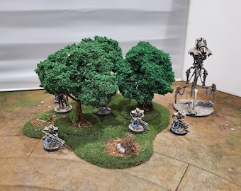 May include: A miniature tabletop gaming scene featuring two green-leafed trees on a grassy island. Several small, detailed figures with weapons and a larger robotic figure are positioned around the trees, suggesting a battle or strategic game.