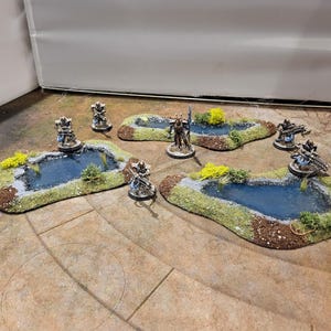 May include: Miniature terrain pieces for tabletop gaming. The set includes several small, irregular-shaped ponds with blue water, green and brown edges, and miniature figures. Yellow and green foliage accents the ponds.