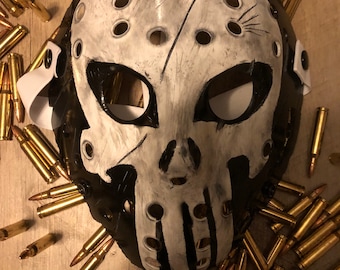 May include: A white and black hockey mask with a skull design. The mask is surrounded by gold-colored bullets.