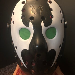 May include: A black and white hockey mask with green mesh eye coverings. The mask has a distinctive design with a white face and black accents. The mask has several small holes and is secured with straps and rivets.