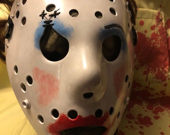 May include: A white hockey mask with red and blue paint, resembling a clown face. The mask has black stitching around the edges and a black strap with a metal buckle.