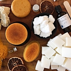 May include: A collection of soap-making supplies including white shea butter chunks, orange circular soap bars, a black bowl of white wax, a bottle labeled Patchouli Oil, yellow turmeric powder, dried orange slices, and wooden stir sticks.