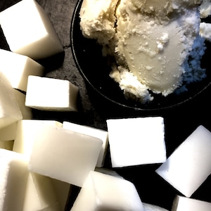 May include: A close-up of a pile of white shea butter blocks, with a bowl of raw shea butter in the background. The text "SHEA BUTTER BASE" is printed on the black surface below the blocks.