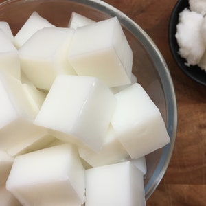May include: A clear glass bowl filled with white, cubed soap bars. The soap bars are arranged in a pile, with some of the bars overlapping. There is a small black bowl filled with white, crumbly soap next to the glass bowl.