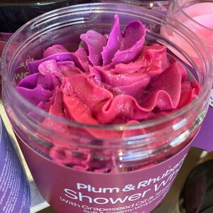 May include: A clear jar with a purple and pink swirl of shower gel inside. The label reads "Plum & Rhubarb Shower Whip with Grapeseed Oil & Soft & Creamy Whipped Soap". The jar is labeled as vegan and cruelty-free.