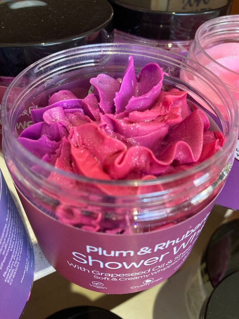 May include: A clear jar with a purple and pink swirl of shower gel inside. The label reads "Plum & Rhubarb Shower Whip with Grapeseed Oil & Soft & Creamy Whipped Soap". The jar is labeled as vegan and cruelty-free.