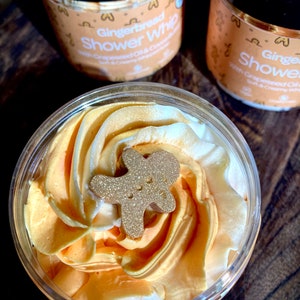 May include: A close-up of a clear jar filled with a golden yellow and white whipped shower cream. A gold glitter gingerbread man is on top of the cream. The jar is labeled "Gingerbread Shower Whip" with "Grapeseed Oil & Coconut"