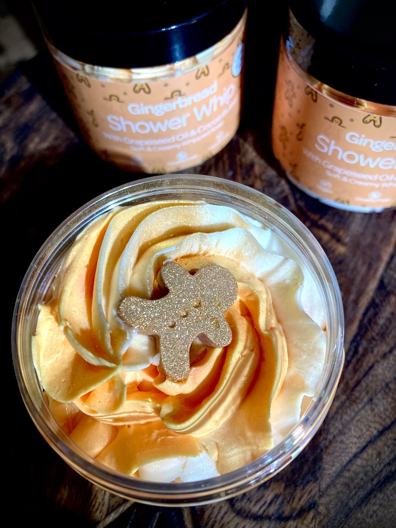 May include: A close-up of a clear jar filled with a golden yellow and white whipped shower cream. A gold glitter gingerbread man is on top of the cream. The jar is labeled "Gingerbread Shower Whip" with "Grapeseed Oil & Coconut"