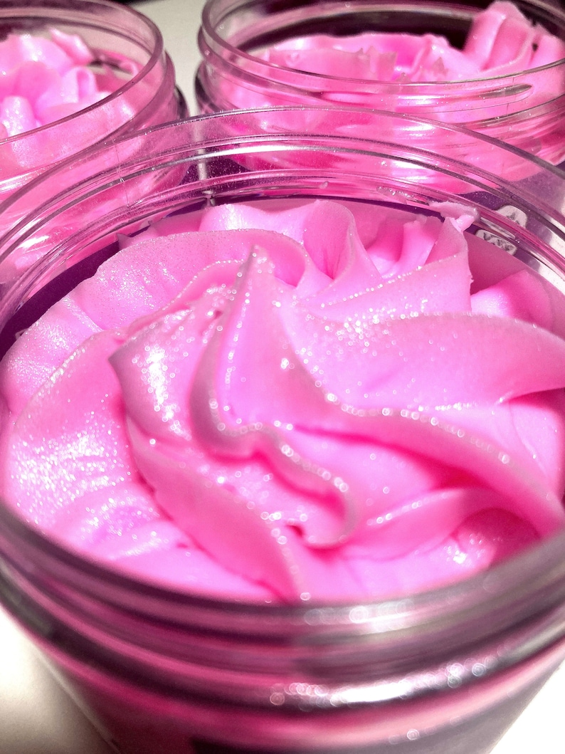 May include: Close-up of a pink whipped body butter in a clear jar with a silver lid. The body butter has a swirl design and a shimmery finish.