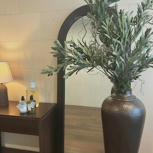 Peut inclure: Un grand vase marron foncé en métal martelé contient des branches d'olivier avec des feuilles vertes et des olives noires. Une table marron foncé avec une lampe et des flacons se trouve à gauche d'un grand miroir en arc. La pièce a une palette de couleurs neutres.
