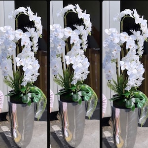 Op de afbeelding: Drie witte orchideeënplanten in zilveren potten. De orchideeën zijn in een vaasvormige vorm met groene bladeren gerangschikt.