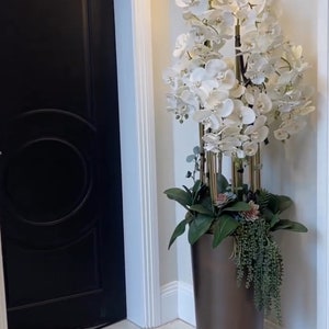 Puede incluir: Una gran planta de orquídea blanca en una maceta de cerámica marrón, colocada en una esquina de una habitación con una puerta negra y paredes blancas.