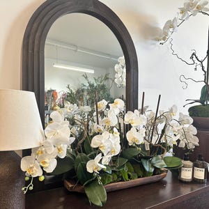 May include: A dark wooden console table with a large, arched mirror. A white lamp with a beige lampshade is on the left. A wooden trough holds white orchids with green leaves and moss. Two small perfume bottles are on the right.