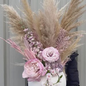 May include: A large, round white vase filled with a floral arrangement. The arrangement includes large pink peonies, lavender roses, and delicate white blossoms, with feathery beige pampas grass and pink accents. The vase is held by a person.